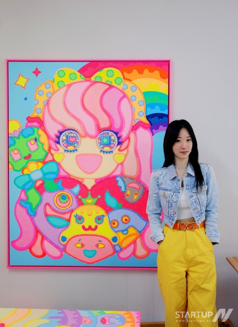 행복과 사랑, 즐거움으로 가득 찬 유토피아를 연상케하는 원더랜드 세계관의 창시자, 이사라 슬롯 사이트가 본인의 작품 앞에서 포즈를 취하고 있다.